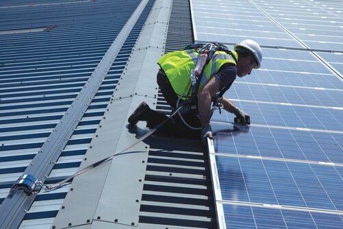 Solar technician wearing a hard hat, high-visibility clothing and fall-protection harness on an industrial roof